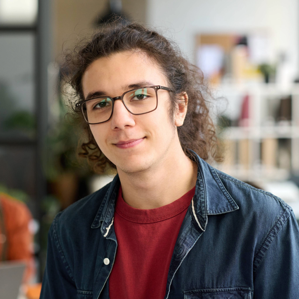A young man with long curly hair tied back, wearing glasses, a red t-shirt, and a denim button-up shirt, smiling slightly while standing indoors in a modern office space.
